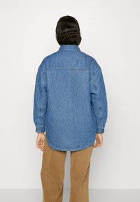 Fodrad denimskjorta i ljusblått, med långa ärmar, krage och en rundad nederkant. På baksidan syns en diamantmönstrad sömnad. Bruna byxor syns.