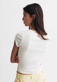 Weißes, kurzärmliges Baumwoll-T-Shirt mit Rundhalsausschnitt und orangefarbenem Logo auf der Rückseite, kombiniert mit gelben Shorts.