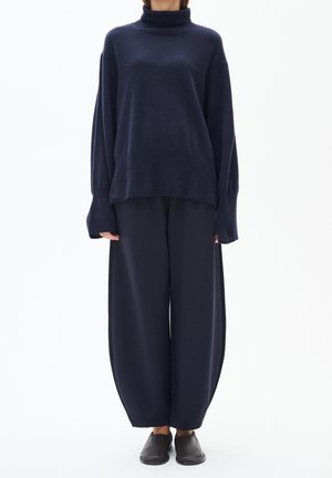 Person iført en løs mørkeblå rullekravesweater, vidde mørkeblå bukser og sorte slip-on sko, stående mod en hvid baggrund.