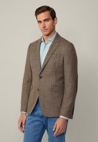 Braun karierter Blazer mit Revers, Knopfverschluss, zwei vorderen Taschen und Ellenbogenpatches, kombiniert mit einem hellblauen Hemd und Jeans.
