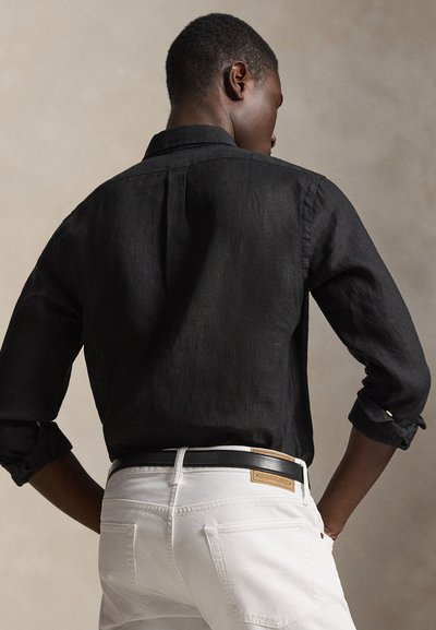 Άνδρας που στέκεται με τα χέρια στις τσέπες, φορώντας μαύρο πουκάμισο με μακριά μανίκια και λευκό παντελόνι με μαύρη ζώνη, γυρισμένος προς την αντίθετη κατεύθυνση από την κάμερα.