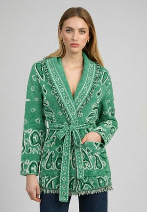 Femme portant un cardigan vert à motifs avec ceinture, détails en franges et boucles d'oreilles créoles dorées, debout devant un fond gris uni.