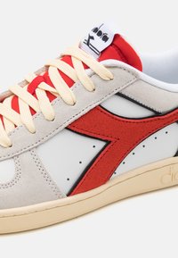 Lærredssneakers med kremfargede og røde detaljer, en grå semsket overdel, hvit perforert panel, tykke snørebånd og en lys tan gummisåle.