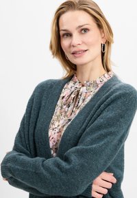 Teal vest met een zachte, gestructureerde afwerking, met een sjawl-kraag en een open voorkant, gelaagd over een delicate blouse met een bloempattern.