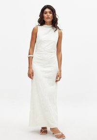 Valkoinen pitkähihaton maxi-mekko, jossa on korkea kaula-aukko, teksturoitu kukkakuosi, vartaloa myötäilevä siluetti ja sileä kangas. Mallilla on päällä valkoiset sandaalit.