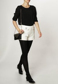 Svart kortärmad topp, vita höga shorts med dekorativ bältesdetalj, svarta strumpbyxor och ankelskots. Modell som håller en liten väska.