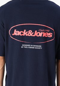 Tums tumši zila kokvilnas T-krekls ar lielu rozā logotipu, uz kura aizmugurē redzams uzraksts "Jack & Jones" un "Club1990", zem tā balti uzraksti.