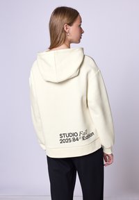 Felpa color crema con cappuccio foderato, caratterizzata da testo nero "STUDIO Fall 2025 (84ª Edizione)" sulla schiena. Vestibilità rilassata e texture morbida.