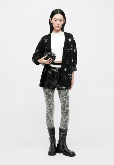 Cardigan negru tricotat cu model de stele, top alb scurt, pantaloni scurți negri din piele ecologică, colanți cu imprimeu de șarpe și cizme negre până la genunchi.