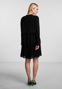 Abito nero a pieghe con un design stratificato e maniche lunghe. Lunghezza al di sopra del ginocchio con una texture fluida. Indossato con scarpe verde con tacco alto.