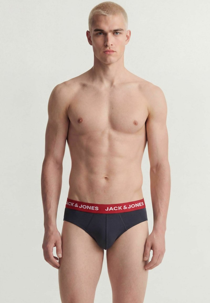 Jeune homme musclé aux cheveux blonds courts portant un slip sombre avec une taille rouge portant l'étiquette Jack & Jones, debout devant un fond uni.