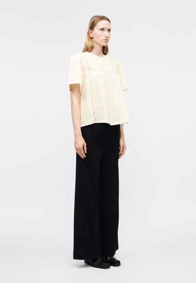 Γυναίκα που φοράει μπλούζα σε μπεζ χρώμα με κοντά μανίκια και διάτρητες λεπτομέρειες, φαρδιά μαύρη παντελόνα και μαύρα παπούτσια, στέκεται μπροστά σε απλό φόντο.