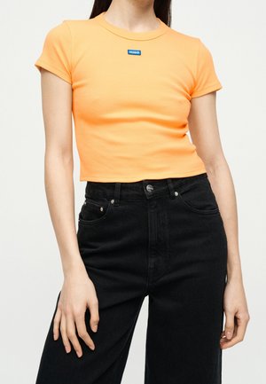 Kobieta nosząca dopasowany pomarańczowy crop top z krótkim rękawem i logo "Hugo" oraz czarne dżinsy z wysokim stanem na jednolitym tle.