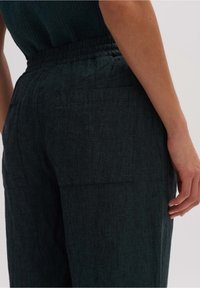 Mörkgröna linnebyxor med elastisk midja och två bakfickor; texturen verkar något grov och andas.