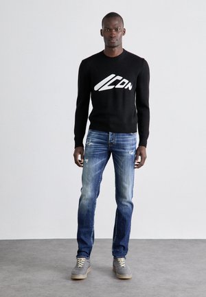 Svart stickad tröja med vitt logo på bröstet, i kombination med slitna blå jeans. Modellen bär grå sneakers med ljusa snören.