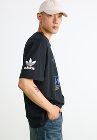 Sort bomulds-t-shirt med korte ærmer, med hvidt Adidas-logo på ærmet og blå grafisk tekst på forsiden. Glat stofstruktur.