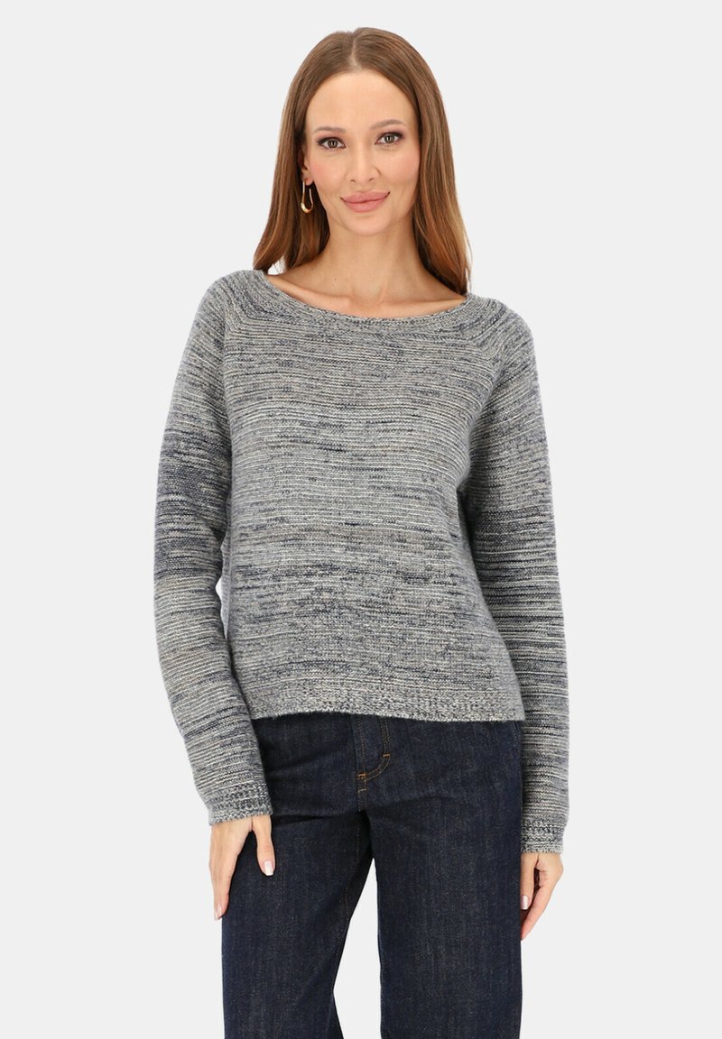 Kobieta o długich brązowych włosach, nosząca złote kołowe kolczyki, szary sweter z dzianiny i ciemnoniebieskie dżinsy, stojąca na jednolitym tle.