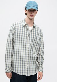 Ung mann med blå baseballcap, hvit og blå rutete skjorte med knapper over hvit t-skjorte, står mot en enkel lys bakgrunn.