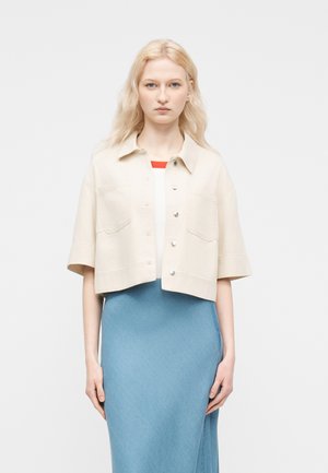 Giovane con lunghi capelli biondi che indossa una giacca corta beige sopra una maglietta bianca e una gonna blu, in piedi davanti a uno sfondo semplice.