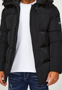 Svart pufferjacka med avtagbar fuskpälsluva, quiltad design, dragkedja framtill och sidofickor. Buren över en vit t-shirt.