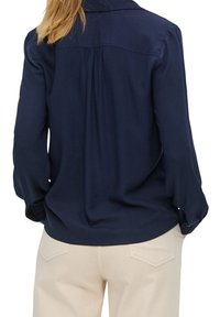 Marineblaue Langarmbluse mit gerafftem Rückenteil und geknöpften Manschetten, kombiniert mit hellbeigen Hosen. Glatte Stofftextur.