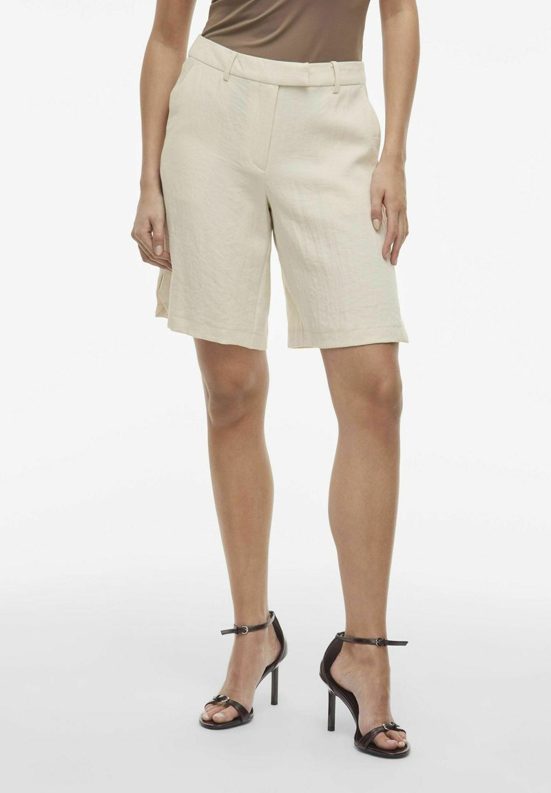 Pantaloni corti di lino beige con una vestibilità rilassata, lunghezza fino a metà coscia, pieghe frontali e passanti per cintura. Abbinati a sandali neri con tacco alto e cinturini.