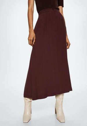 Vrouw draagt een asymmetrische midi-rok in donkerbruin met hoge taille en witte laarzen met hak, staande tegen een effen achtergrond.