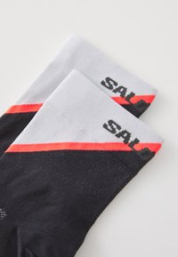 Calcetines deportivos en negro y gris con un diseño texturizado, adornados con una franja de acento roja y el logotipo "SAL" impreso cerca del borde superior.