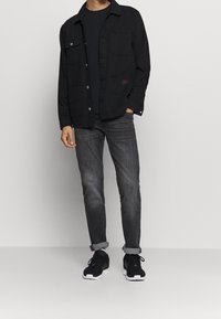 Hombre con chaqueta negra sobre camisa negra, jeans gris deslavado enrollados en los puños y zapatillas negras con suelas blancas, de pie sobre un fondo liso.