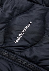 Svart quiltad väst med dragkedja och "Peak Performance"-logotyp i vit brodering på högra sidan. Slät textur, lätt material.