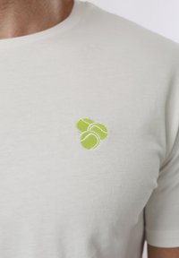 Κοντινό πλάνο λευκού μπλουζακιού με μικρό κεντητό σχέδιο τριών επικαλυπτόμενων πράσινων τενιστικών μπαλών στο στήθος.