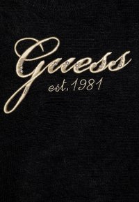 Il tessuto nero presenta la scritta ricamata "Guess" in oro con perle decorative, accanto alla dicitura "est. 1981" in un carattere più piccolo.