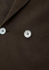 Brązowa marynarka z tkaniny z dwoma widocznymi okrągłymi białymi guzikami z czterema dziurkami, pokazująca klapy i szczegóły tekstury z bliska.