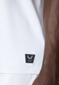Weißes Sportshirt aus strukturiertem Stoff mit einem kleinen schwarzen rechteckigen Etikett mit Flügelmuster und dem Text "AUTHENTIC CASTORE PRODUCT".