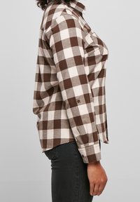 Femme portant une chemise à manches longues à carreaux marron et blanc et un pantalon noir, debout de profil devant un fond clair uni.