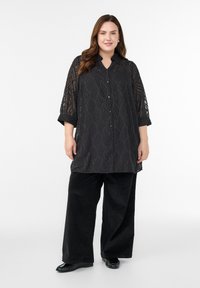 Žena, ktorá má na sebe čiernu priesvitnú vzorovanú tuniku na gombíky s vyhrnutými rukávmi, čierne široké nohavice a čierne balerínky, stojaca na bielom pozadí.