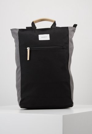 Schwarzer und grauer Sandqvist-Rucksack mit beigem Tragegriff oben und Frontreißverschlusstasche auf einem weißen Podest.