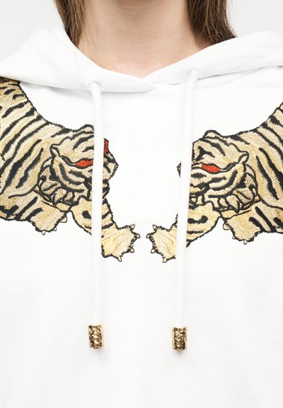 Λευκό φούτερ με κουκούλα και κεντημένους συμμετρικούς τίγρεις που κοιτάζουν ο ένας τον άλλον στο ύψος του στήθους, με κόκκινα μάτια και λεπτομέρειες ραβδώσεων στο τρίχωμα.
