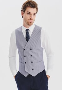 Jasnoszara kamizelka w paski z podwójnym zapięciem na guziki, klapami typu notch, w połączeniu z białą koszulą i ciemnym wzorzystym krawatem.