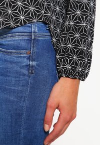 Svart topp med vitt geometriskt mönster, i kombination med blå denimjeans. Tyget har en mjuk textur; jeansen har klassisk femficksdesign.