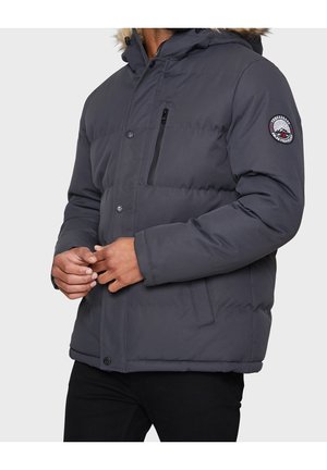 Homme portant une veste d'hiver matelassée gris foncé avec une capuche bordée de fourrure et un écusson sur la manche gauche, en train de fermer les boutons devant.