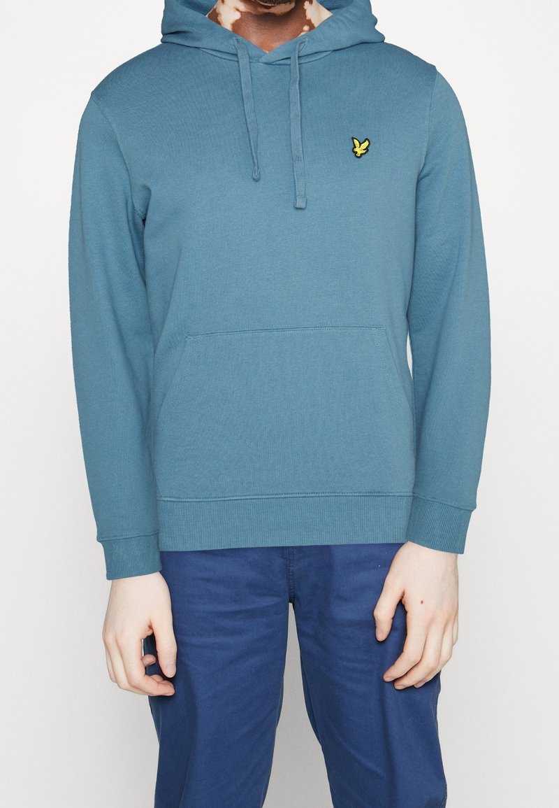 Personne portant un sweat à capuche bleu sarcelle avec un logo d'oiseau jaune sur la poitrine et un pantalon bleu, debout devant un fond blanc uni.
