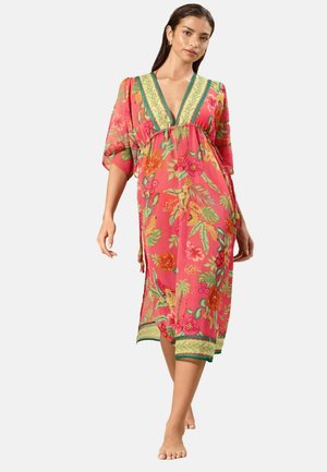 Frau, die barfuß ein rosa geblümtes Midikleid mit grünem und gelbem Rand und tiefem V-Ausschnitt vor einem schlichten Hintergrund trägt.