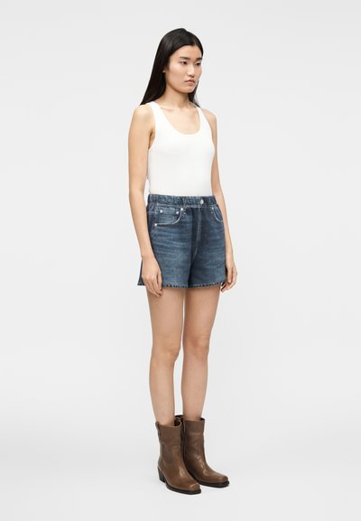 Shorts di jeans con lavaggio blu scuro, caratterizzati da una vita aderente con passanti per cintura. Abbinati a una canotta bianca a coste e stivaletti marroni alla caviglia.