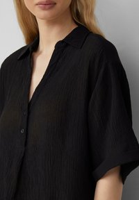 Schwarzes Text-Shirt mit Kragen, kurzen Ärmeln und Knopfleiste, aus einem leichten, zerknitterten Stoff gefertigt.