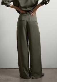 Personne avec les mains sur le bas du dos portant un pantalon large olive vert loose et un haut assorti devant un mur clair.