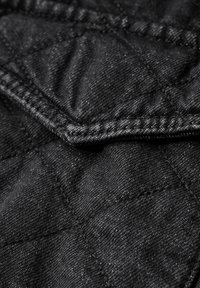 Tela negra acolchada con un acabado texturizado. Presenta costuras diagonales y una costura doblada, resaltando un diseño duradero.