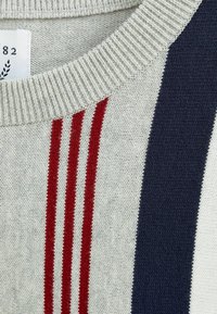 Pull en maille gris clair avec encolure côtelée. Présente des rayures verticales rouge et bleu marine. La texture semble douce et légèrement ajourée.