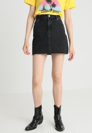 Frau trägt einen schwarzen Denim-Minirock mit ausgefranstem Saum, ein gelbes T-Shirt mit Grafik und schwarze glänzende Stiefeletten, steht mit entspannten Armen.