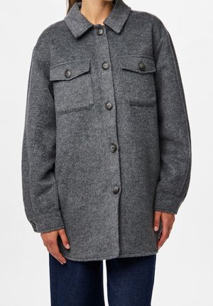 Nainen pukeutunut löysään, pitkähihaiseen harmaaseen villatakkiin, jossa on napit, kaksi rintataskua ja tummansiniset farkut.